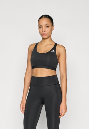 Sujetador deportivo negro con tirantes anchos y escote redondeado, con un logo blanco en el lado izquierdo. Tela lisa y elástica. Combinado con leggings negros de cintura alta.
