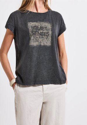 Kobieta ubrana w wyblakły czarny t-shirt z nadrukiem "QUIET SENSES" w surowym kwadracie i beżowe spodnie, ręce w kieszeniach, na białym tle.
