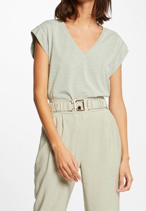 Kobieta ubrana w jasnozielony, bezrękawnik z dekoltem w kształcie litery V, włożony w beżowe spodnie z wysokim stanem, ozdobione szerokim materiałowym paskiem z kwadratową klamrą.