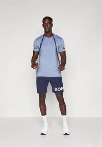 Lys blå kortermet skjorte med "BORG"-logo, kombinert med marineblå shorts med fet hvit "BORG"-tekst. Modellen holder en vannflaske.