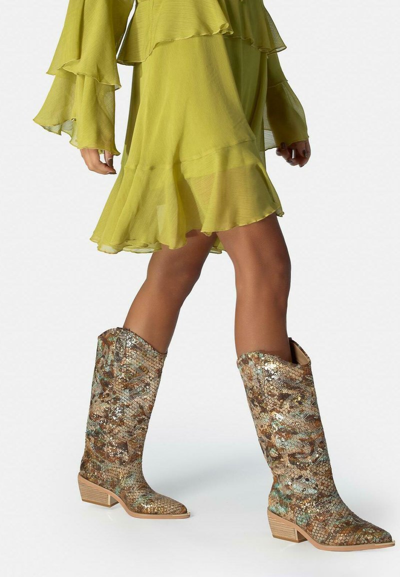 Personne marchant dans une robe vert citron mi-longue et transparente, avec des bottes montantes à motifs peau de serpent et talons bas.