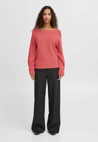 Koralen pleteni pulover s širokim izrezom in dolgimi rokavi, kombiniran s črnimi širokimi hlačami. Gladka tekstura in sproščen kroj.