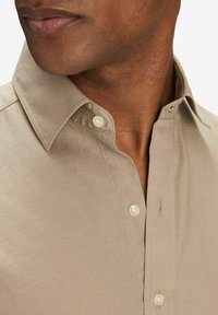 Beige skjorta med knappar och spetsig krage, som har två knappar framtill och en slät tygtextur. Närbild med fokus på halslinningen.