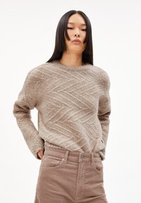 Frau mit glattem, dunklem Haar trägt einen strukturierten beigen Pullover und hochtaillierte braune Cordhose, steht vor einem weißen Hintergrund.