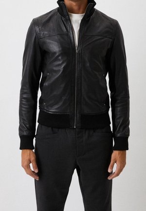 Mann in einer schwarzen Lederjacke mit Reißverschluss, gerippten Bündchen und Saum, über einem weißen Hemd und schwarzen Hosen, stehend vor einem schlichten Hintergrund.