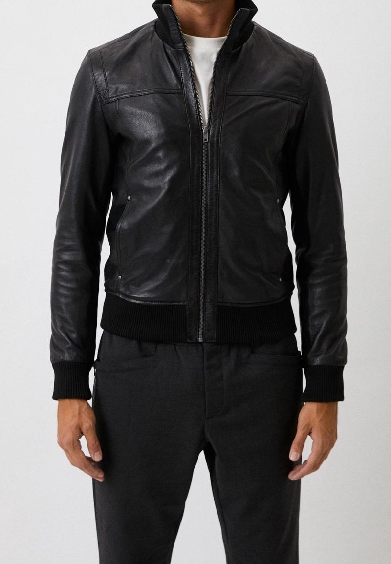 Mężczyzna w czarnej skórzanej kurtce zapinanej na zamek z żebrowanymi mankietami i dołem, noszący białą koszulę i czarne spodnie, stojący na jednolitym tle.