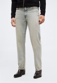 Άνδρας που φοράει τζιν με ίσια γραμμή σε ανοιχτό πλύσιμο, μαύρο μπουφάν με φερμουάρ και μαύρα δερμάτινα μποτάκια αστραγάλου, στέκεται μπροστά σε απλό φόντο.