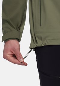 Vandafvisende grøn jakke med elastiske manchetter og justerbar kant. Håndtag til at trække snoren op fra bunden. Glat, letvægtsstof.