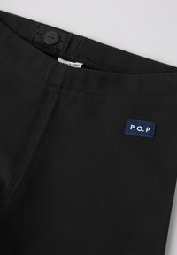 Mustat puuvillahousut, joissa on sileä pinta, joustava vyötärö, jossa on nappikiinnitys, ja sininen "P.O.P" -tägki ommeltuna etuosaan.