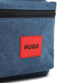 Denim bag with a textured blue exterior, featuring a black zipper and a red rectangular patch with the word "HUGO" in bold black letters.
