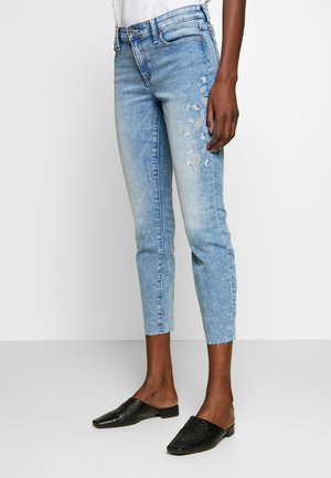 Vaqueros de mezclilla azul claro con un ajuste Slim, de longitud recortada y un detalle de bordado en paisley blanco en un lado. Combina con zapatos negros de textura tipo slip-on.