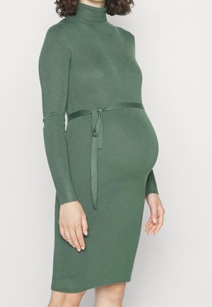 Femme enceinte portant une robe en maille verte ajustée à manches longues avec un ruban noué autour de la taille sur un fond uni.