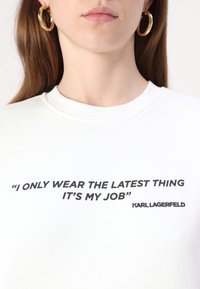 Felpa bianca con una citazione nera stampata sul davanti: "INDOSSO SOLO L'ULTIMA NOVITÀ, È IL MIO LAVORO" con il logo "KARL LAGERFELD".