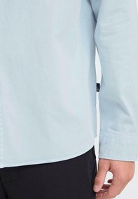 Lichtblauwe blouse met lange mouwen met een zachte textuur, knoopmanchetten en een afgeronde zoom, met een label zichtbaar nabij de naad.