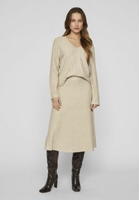 Beiges gestricktes Zweiteiler-Outfit mit einem gerippten Langarmoberteil mit V-Ausschnitt und einem passenden knielangen Rock. Stilisiert mit glänzenden kniefreien Stiefeln.