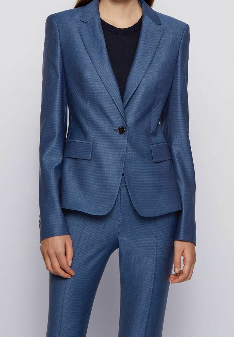 Blazer bleu sur mesure en tissu texturé, avec une fermeture à un bouton, des revers à encoche et deux poches avant. Associé à un haut noir.
