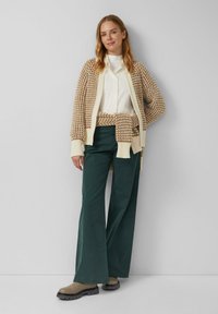 Strickcardigan im beigefarbenen und braunen Muster, weißes Kragenshirt, türkisfarbene weit geschnittene Hose und braune Ankle Boots. Sanfte Texturen und entspannter Schnitt.