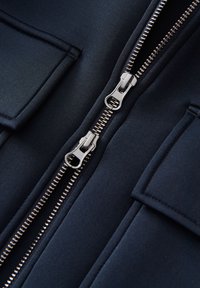 Giacca blu navy con doppi zip argento, tasche piatte e una texture di tessuto liscia, caratterizzata da linee pulite ed elementi di design minimalisti.