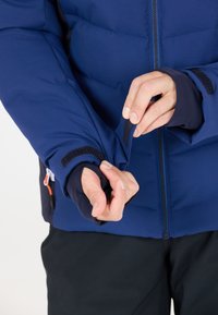 Blå isolerad jacka med dragkedja fram och justerbara ärmändar, med en slät textur och elastiskt inre lager för komfort.