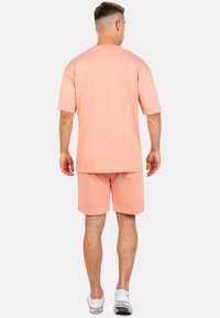 T-shirt oversized di cotone color pesca con maniche corte, abbinato a pantaloni coriaceo coordinati. I pantaloni presentano una tasca posteriore e una vestibilità rilassata.