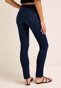 Jeans ajustados de color azul oscuro con una cintura alta, que cuentan con dos bolsillos traseros y una textura suave, combinados con sandalias negras de tiras.