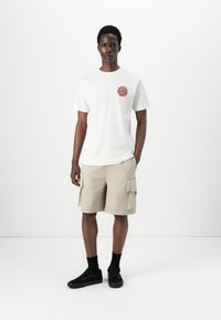 White t-shirt with a red circle logo that says "Santa Cruz," paired with beige cargo shorts and black sneakers. Minimalist design, casual fit.