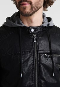 Veste en simili cuir noir avec une capuche grise, fermeture éclair à l'avant, accents en métal argenté et tissu texturé, offrant une coupe décontractée.