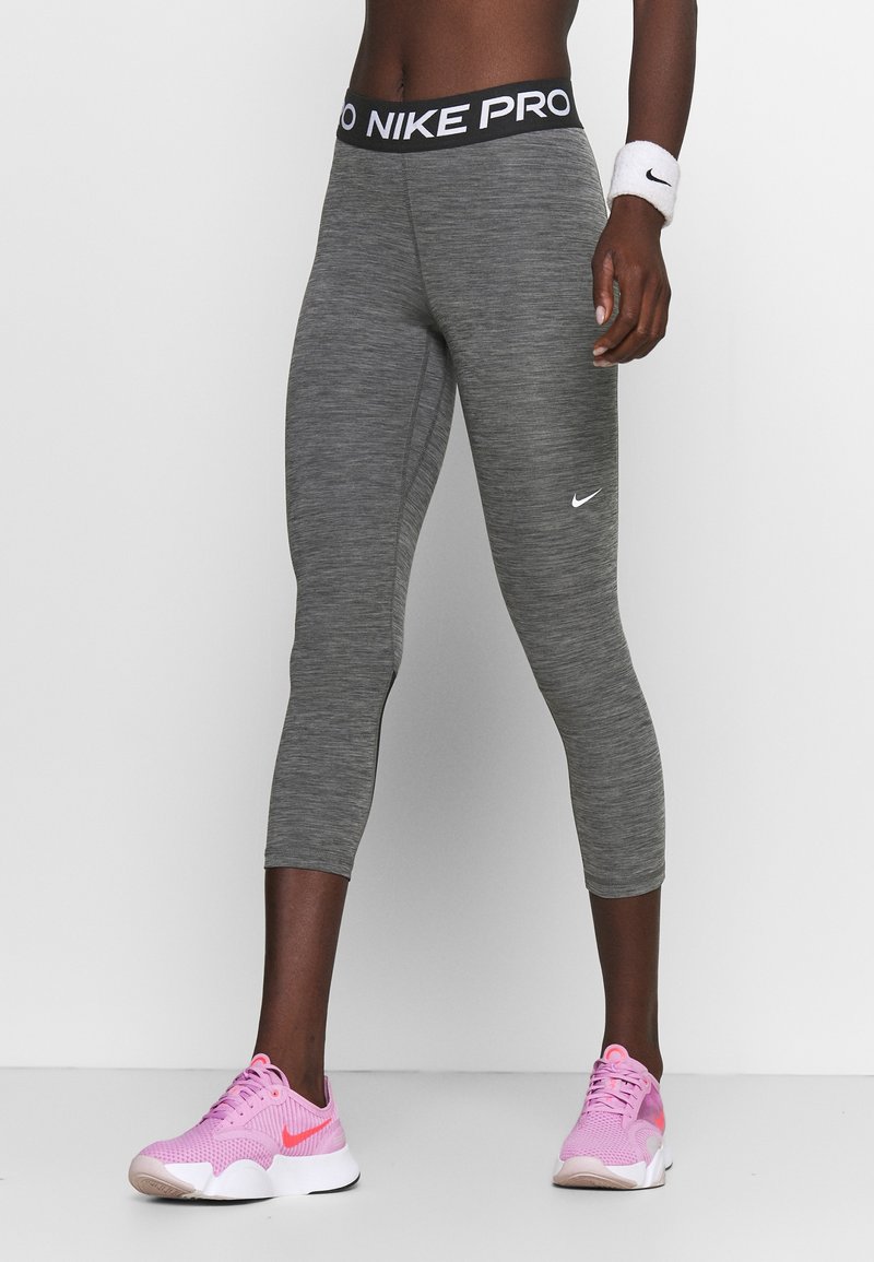 Personne portant des leggings gris coupés Nike Pro, des baskets roses Nike et un bracelet Nike blanc, debout devant un fond uni.
