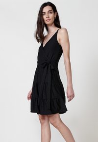 Schwarzes Wickelkleid mit stufenförmigem Design, aus strukturiertem Stoff gefertigt. Mit verstellbaren Trägern und einem gebundenen Taillendetail.