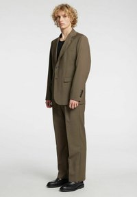 Brūna, pēc pasūtījuma šūta uzvalka, kas sastāv no vienas pogas žaketa, atbilstošiem biksēm un melniem apaviem. Gluda auduma ar strukturētu piegriezumu.