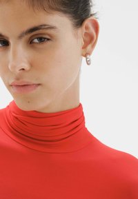 Pendientes de aro de plata con una superficie texturizada y un corte en forma de corazón, combinados con una blusa roja ajustada de cuello alto.