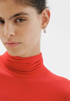 Pendientes de aro de plata con una superficie texturizada y un corte en forma de corazón, combinados con una blusa roja ajustada de cuello alto.