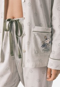 Set di pigiama in cotone con una base grigia chiara e motivi di pois e fiori verdi. Include una camicia con bottoni, pantaloni con cordoncino e una tasca con ricamo.