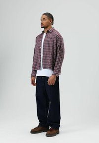 Kariertes Hemd mit Knöpfen in Rot und Grau, über einem weißen T-Shirt getragen, kombiniert mit dunklen Jeans und braunen Schnürstiefeln.