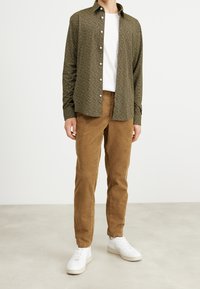 Camicia verde a fantasia con bottoni, indossata sopra una t-shirt bianca. Pantaloni in velluto a coste marroni e sneakers bianche completano l'outfit.