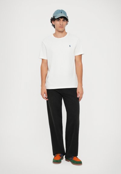 T-shirt branco com um pequeno logótipo azul, calças largas pretas e um boné azul claro. Sapatos multicoloridos com detalhes laranja e verde.