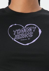 Černé bavlněné tričko s fialovým srdcovým designem a textem "TOMMY JEANS" a "Since 85" tučným písmem.