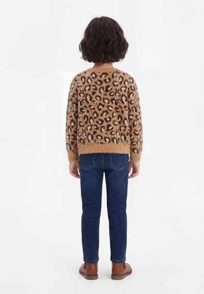Dieťa s kučeravými tmavými vlasmi otočené chrbtom, oblečené v huňatom béžovom svetri so vzorom leopardieho vzoru, modré džínsy a hnedé topánky na bielom pozadí.