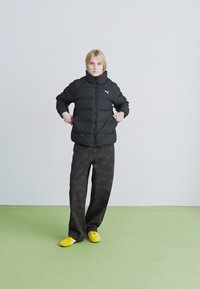 Szőke személy fekete Puma steppelt kabátban, sötét mintás bőrszárú nadrágban és élénk sárga sportcipőben áll egy zöld padlón.