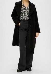Cappotto di lana nero con colletto, abbinato a una blusa a stampa zebra bianco e nero e jeans a vita alta neri a gamba larga. Scarpe nere con suola in gomma.