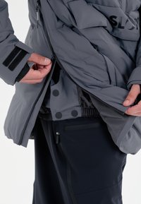 Grå isoleret jakke med lynlås og trykknaplukninger. Har velcro-manchetter og en snøret talje. Bæres over sorte bukser.