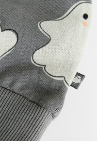Harmaa neule, jossa valkoinen kummituskuvio; ribbauskudoksen rakenne; sisältää pienen etiketin, jossa on kummituskuva. Pehmeä neulekangas.