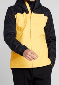 Žlutá a černá voděodolná bunda s kapucí, elastickými manžetami a přední kapsou. Hladký povrch, lehký design.