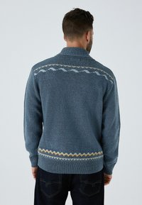 Férfi kék kötött pulóver magas nyakkal; fehér, sárga és kék mintázott díszítések találhatók a vállakon és a szegélyen. Texturált anyag.