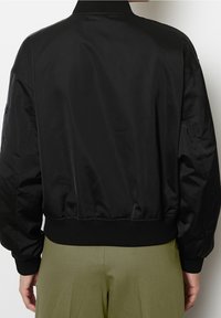 Chaqueta bomber negra con una textura lisa y brillante, puños y dobladillo acanalados, y un ajuste relajado. Costuras visibles realzan el diseño.