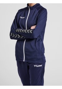 Mörkblå zip-up jacka med texturerad tyg, med vit dragkedja och grå chevronaccenter på ärmarna. Kombineras med matchande byxor.