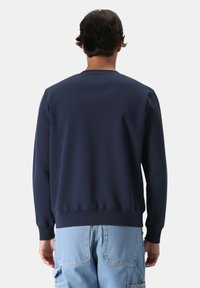 Uomo con capelli scuri e corti, indossante un maglione blu navy e pantaloni cargo in denim azzurro chiaro, di spalle su uno sfondo neutro.