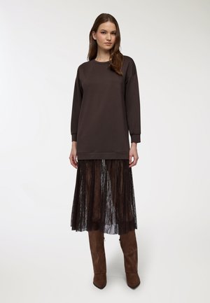 Sweatshirt oversize marrone scuro con maniche lunghe, indossato sopra una gonna in pizzo nero trasparente. Presenta un collo rotondo e un orlo a coste.
