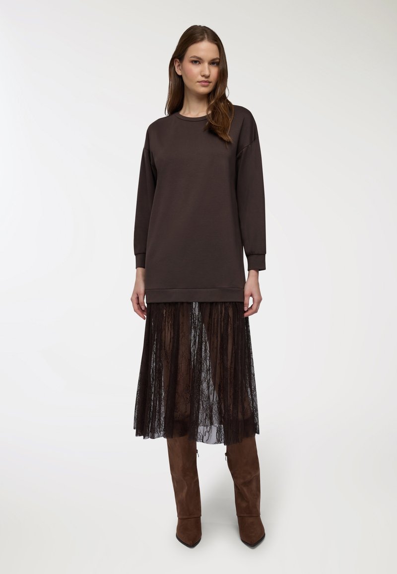Sweatshirt oversize marrone scuro con maniche lunghe, indossato sopra una gonna in pizzo nero trasparente. Presenta un collo rotondo e un orlo a coste.