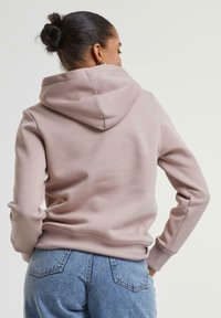 Un hoodie rose clair avec une capuche et des poignets côtelés, associé à un jean bleu taille haute. Le tissu semble doux et légèrement texturé.
